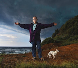 Man with outstretched arms and small dog on a cliff overlooking the ocean.